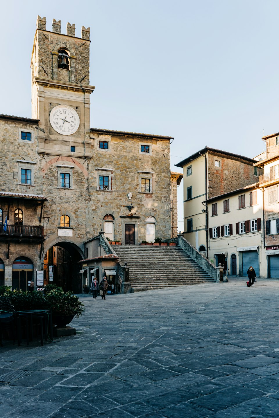 Piazza italiana illuminata dal sole con torre dell'orologio e palazzi in pietra.