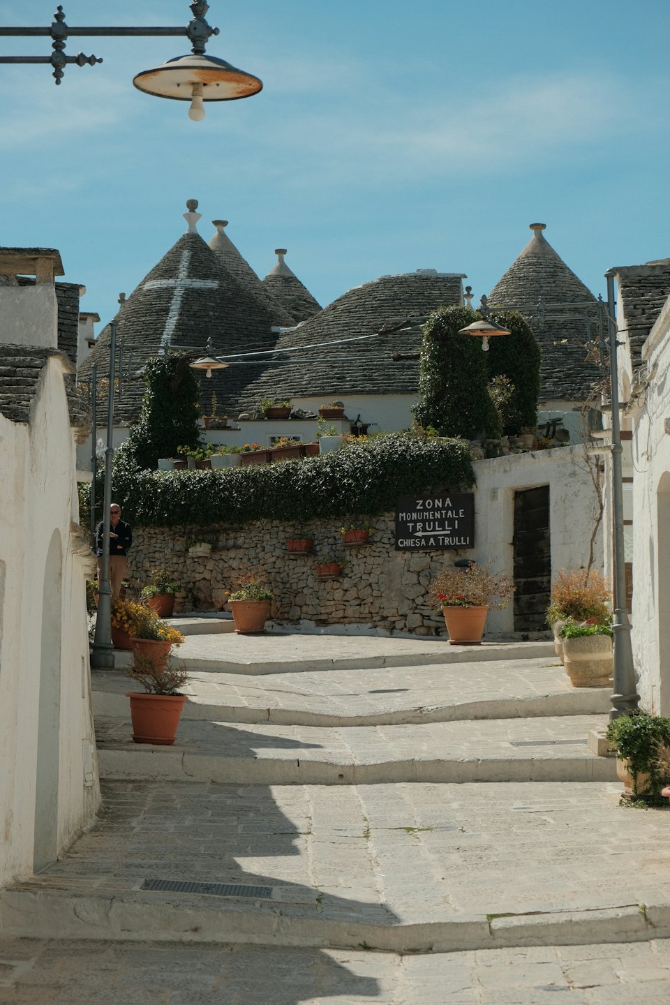 Strada in salita fiancheggiata da trulli bianchi e piante in vaso.